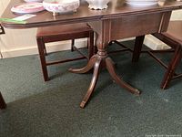 Side view of the wooden drop leaf table showing the table surface, side chairs, and the carved pedestal leg ending in claw feet with brass caps