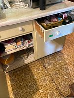 Photo showing underside cabinet with drawers and shelves containing folded kitchen cloths, a yellow storage bowl, glass items, and utensils in the open drawer.