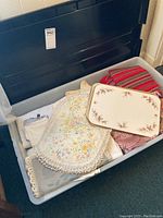 Assortment of linens, lace doilies, embroidered cloth, folded napkins, and decorative serving tray stacked in plastic bin