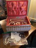 Wicker sewing kit open showing pearl necklaces and wristbands inside, plastic bags containing garnet necklace and metal wristband outside kit