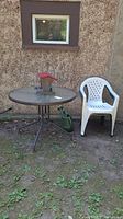 Photo showing the round metal table with glass top, one white PVC chair, green watering can, and red/metal bird feeder on the table.