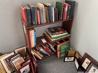 Front view of small wooden bookshelf filled with books and surrounded by picture frames on floor.