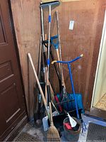 Various outdoor tools including shovels, rakes, broom, dustpan, and bucket grouped together in a corner.