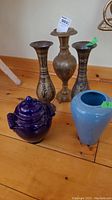 Photo showing three brass vases, one blue ceramic vase, and one blue ceramic lidded bowl on wooden surface.