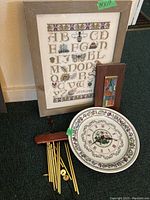 Photo showing framed embroidered alphabet sampler, small framed colorful letter sign, decorative plate clock with floral and butterfly motifs, and wooden wind chime with brass tubes.