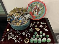 Full view of vintage jewelry collection with jade pendants arranged on a dark cloth next to a decorative metal tin, various brooches and earrings surrounding them.
