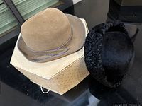 Photo showing beige Louise Green hat and black Francette original hat on a light brown box, both hats displayed on a dark surface.