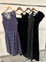 Four vintage dresses hung on a rack showing front and sides. Purple lace sleeveless dress, two black sleeveless velvet dresses, one with floral pattern, one plain black.
