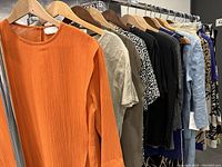 Rack of assorted blouses and tops in multiple colors including bright orange, black, patterned, and beige.