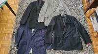 Four men's jackets laid out on a carpet showing various colors and styles including black, gray pinstripe, and navy blue.