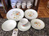 Dishes including dinner, appetizer, bread and butter plates, cups, and saucers arranged on carpet in front of shelf