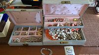 Two gray jewelry boxes open showing assorted costume jewelry inside such as beaded necklaces and brooches, displayed on a table.