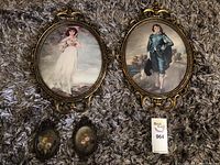 Two large oval metal-framed prints with convex lenses depicting historical figures, a woman and a man, with metal frames and felt backing, shown on carpet.