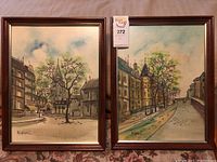 Photo showing both framed artworks side by side on table, two signed canvases depicting European street scenes