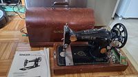 Vintage Singer sewing machine with attached wooden base and brown carrying case in background, manual on left, small accessories to right of machine.