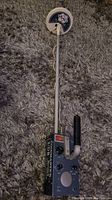 Full view of the metal detector showing white shaft, circular search coil, handle, and control box on carpet.