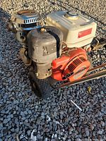 Front angle view showing the Honda GX390MA engine, white fuel tank, black air filter housing, metal frame, and wheel on gravel ground