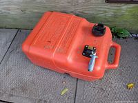 Orange plastic boat fuel tank laying on outdoor surface showing built-in handle, fuel cap, and fuel outlet fitting with attached valve.