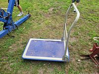 Foldable push cart standing upright on grass with blue flat deck platform and beige handle showing some surface wear.