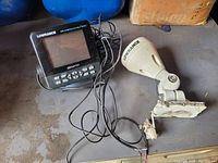 Front view of Lowrance GPS marine unit with screen and control buttons, wiring visible, placed on a floor.