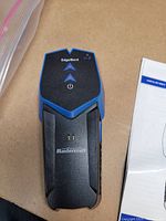 Top view of Mastercraft stud finder with edge detection buttons and LED indicators on black and blue plastic body, alongside user manual paper.