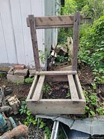 Wooden outboard boat engine stand with wheels placed outdoors near a shed. The stand is constructed from thick, weathered wood with a rectangular base and vertical support beams.