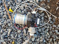 Top view of the RV water pump showing motor label, wiring, and tubing on gravel surface