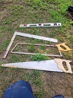 Photo showing two saws, a carpenter's square, and a metal level on grass ground. The items appear used and untested.
