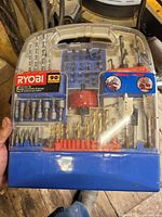 Ryobi branded 90-piece drill bit and screwdriver bit set displayed in clear plastic case with blue base, showing assortment of metal bits organized inside