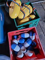 Yellow plastic water floats with rainbow-colored straps stored in a green milk crate. Blue and white oval rope floats in a red milk crate beside it.