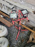 Two-headed portable work light with rectangular lamps on a red adjustable tripod stand placed on gravel and leaning against a wooden wall.