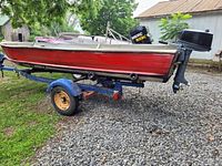 Side view of the red aluminum boat mounted on a blue single-axle trailer, showing dents and outboard motor.