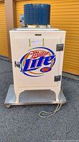 Front view of restored cream-colored fridge on wheeled dolly with hand-painted Miller Lite logo and top compressor unit