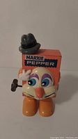 Front view of Marxie Pepper wind-up toy showing the orange box body with the MARXIE PEPPER label, cartoon face, hat, and wind-up key on the side