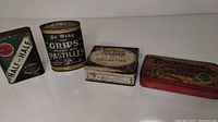 Four antique tins arranged side by side: Burley and Bright Half and Half tin, Grips First Aid Pastilles tin, British Console Cigarettes tin, and a red tin with German text.