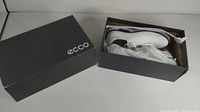 Photo showing the ECCO shoe box with the lid removed and one pair of white golf shoes inside, wrapped partially in tissue paper.
