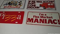 Photo showing four novelty license plate covers with various designs: Walt Disney World, van with phrase, pizza text, flea market mania text.
