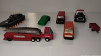 Seven toy vehicles displayed in a row including a fire truck with ladder, toy car, van, police car, mini trailer, and boat