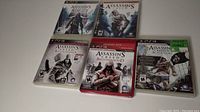 Photo showing six PlayStation 3 Assassin's Creed game cases arranged on a white surface, displaying cover art for Brotherhood, Revelations, Rogue, Black Flag, and two original Assassin's Creed titles.