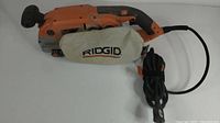 Front angled view of the Ridgid belt sander showing the dust collection bag, power cord, and ergonomic handle.