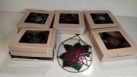 One handmade stained glass poinsettia ornament placed in front of five boxed star-shaped colored glass candle holders in various colors including blue, purple, and red.