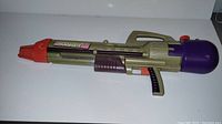 Side view of the vintage Larami Super Soaker CPS 2000 water toy showing the full length of the gun, olive green body with purple tank and orange nozzle.