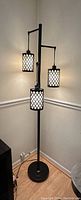 Full view of the black metal floor lamp with three lights, each covered by a lattice patterned cylindrical shade, placed in a corner with wooden floor and beige walls.