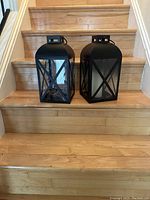 Two black metal lanterns placed on wooden stairs. One lantern contains a white votive candle, the other has battery string lights inside visible through frosted glass. Both have X-shaped front glass panel designs and small top handles.