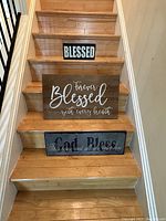 Photo showing three Blessed-themed decorative signs placed on wooden stairs. Two smaller signs on different steps and one larger wooden sign in middle step.