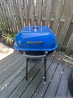 Bold blue Americana charcoal grill with dome lid, placed on wooden deck outside near wooden fence.