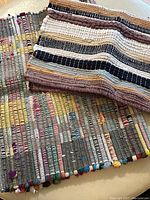 Close-up photo showing two multicolored cotton rag rugs with woven stripes in various colors including black, brown, white, red, yellow, and blue.