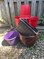 Photo showing three large red plastic flower pots, one purple plastic pot, one brown ribbed plastic pot, and one large red clay pot stacked together outdoors on gravel near a wooden fence.