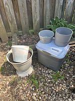 Photo of three grey flower pots and one mid-size grey cooler placed on rocky ground near wooden fence.