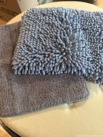 Two grey bath mats on a light surface, showing different pile textures; one with thick microfiber tufts and the other with a flat plush surface.